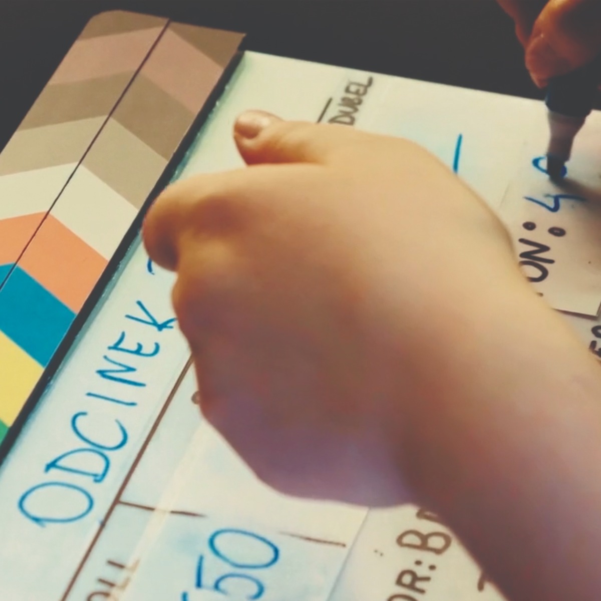 Osoba wypełnia klaps filmowy, na którym widoczny jest napis 'ODCINEK 150' i inne dane dotyczące produkcji filmowej.