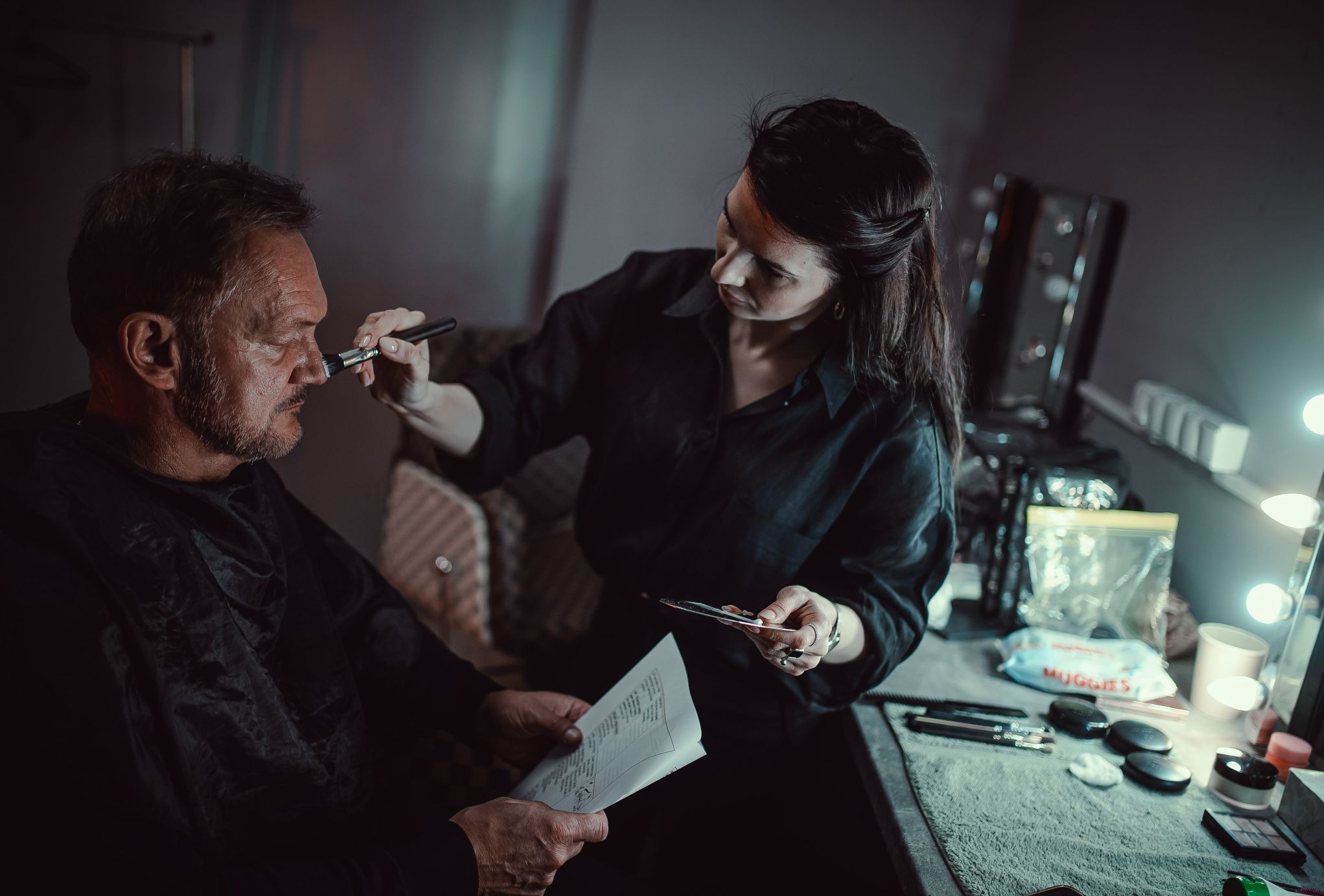 Kobieta nakłada makijaż pędzlem na twarz mężczyzny, który trzyma scenariusz w dłoniach; w tle toaletka z oświetleniem i kosmetykami.