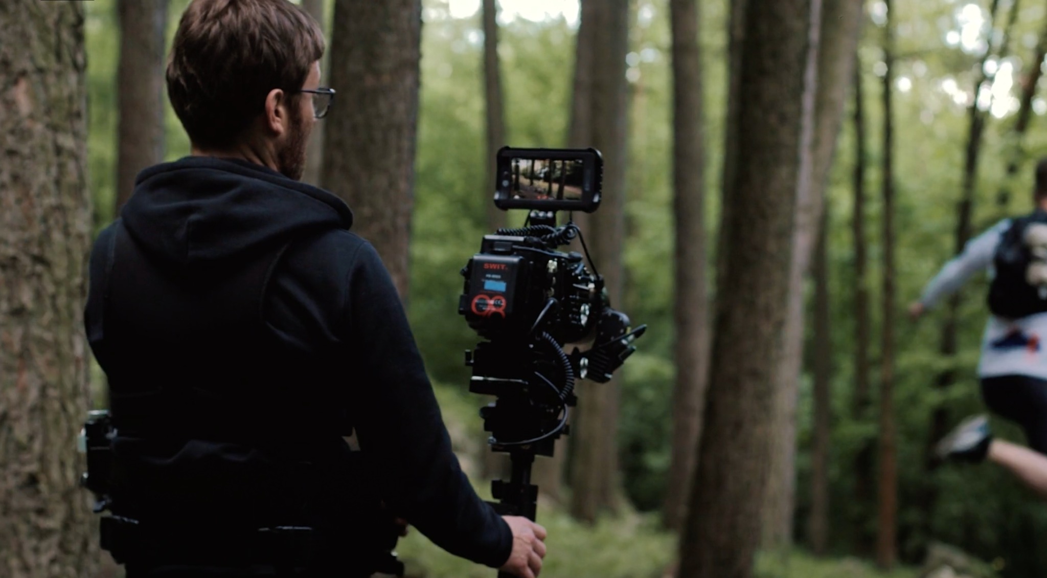 Operator kamery z profesjonalnym sprzętem filmuje biegacza w lesie, widok zza pleców operatora.
