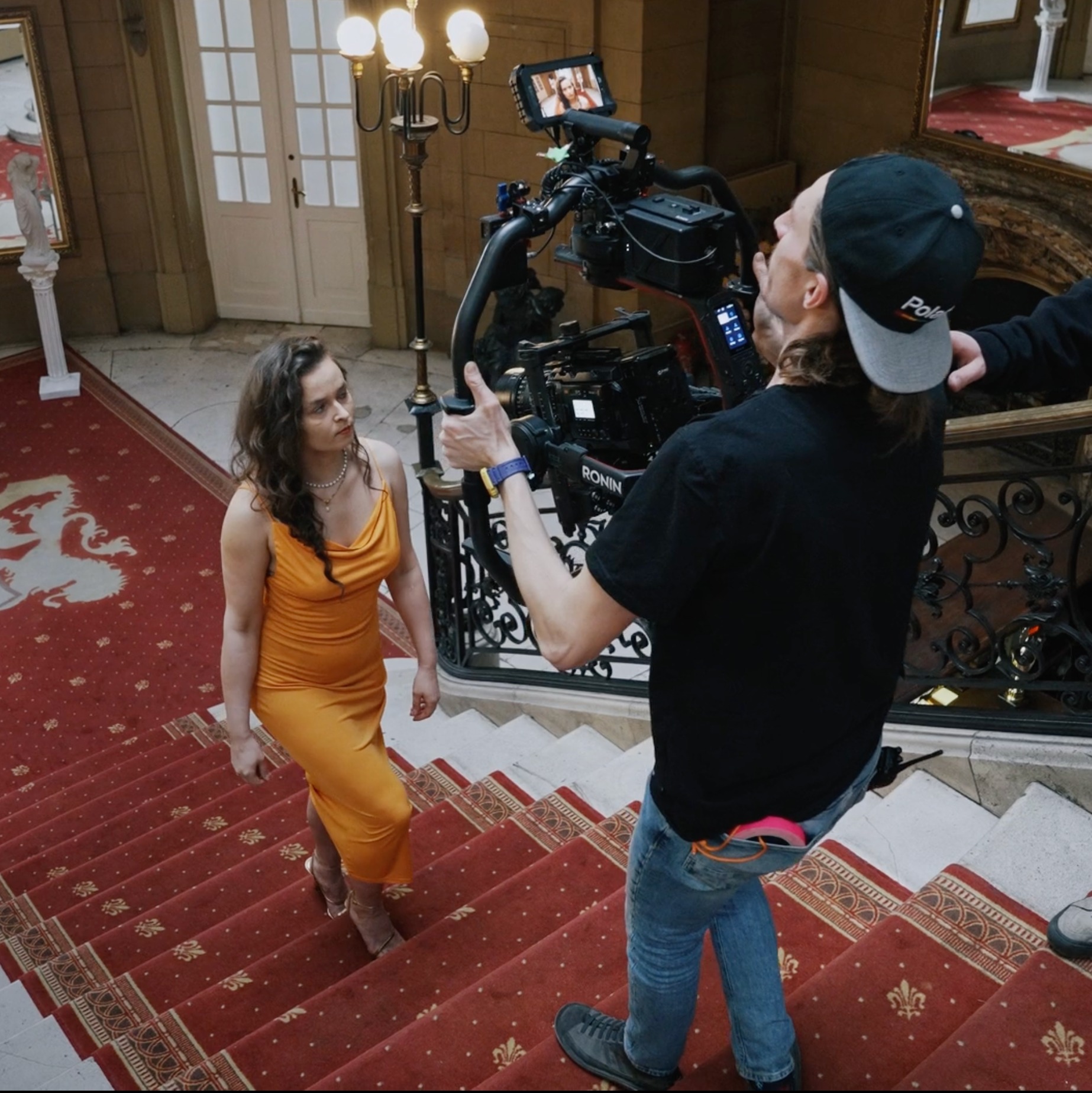 Kobieta w pomarańczowej sukience schodzi po schodach, filmowana profesjonalną kamerą stabilizowaną gimbalem, w tle bogato zdobione wnętrze z czerwonym dywanem i żyrandolem.