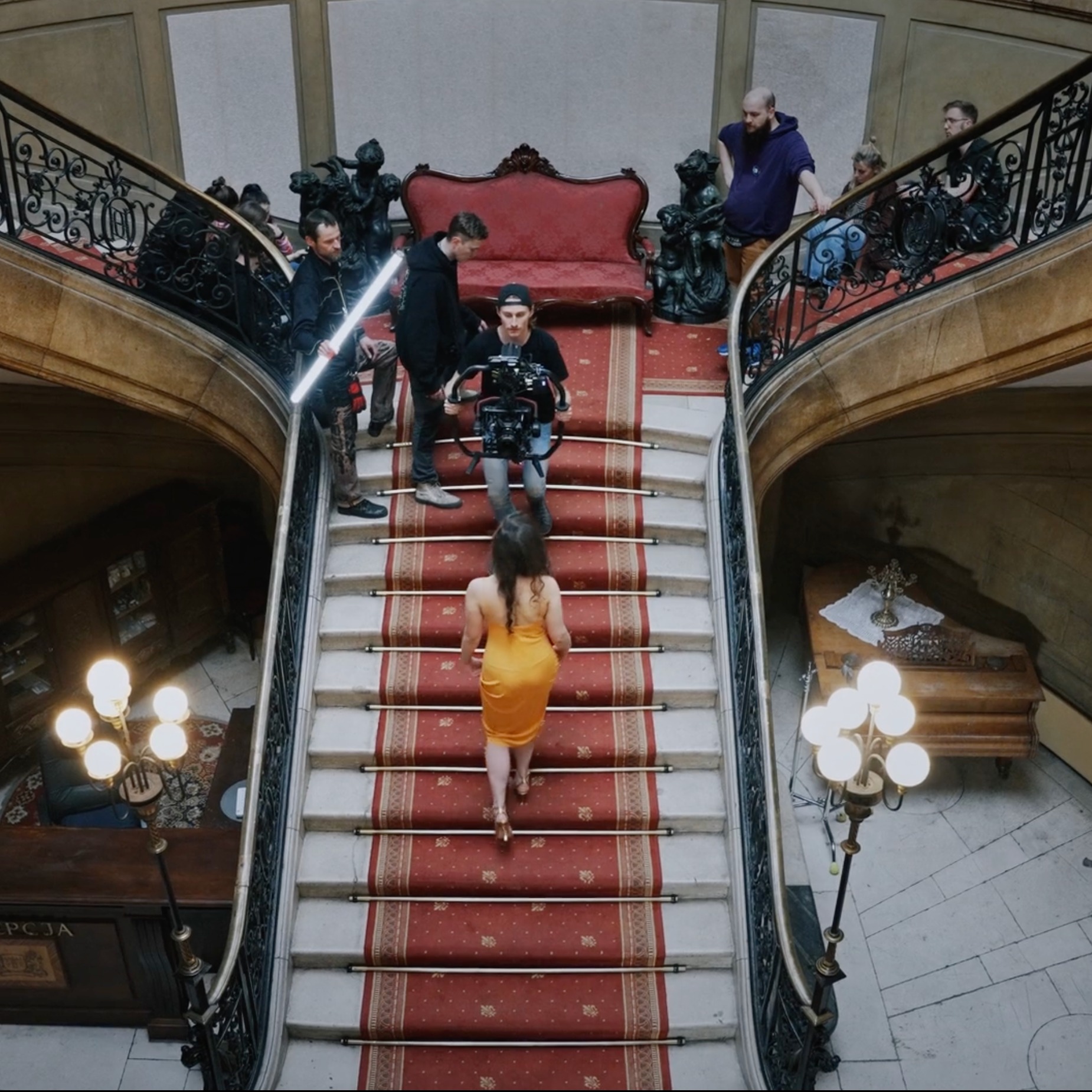 Ekipa filmowa kręci scenę z kobietą w żółtej sukience wchodzącą po schodach w eleganckim wnętrzu, operator z kamerą na stabilizatorze, oświetleniowiec z ledową lampą, rekwizyty w tle.