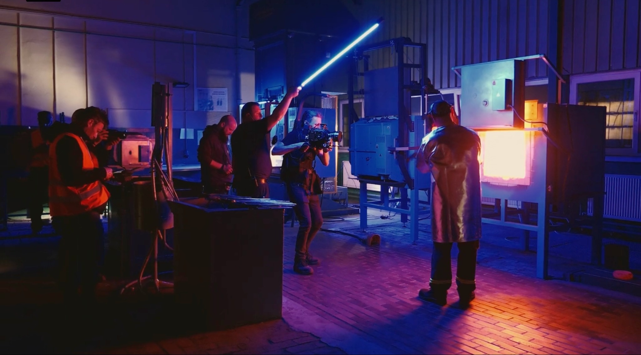 Ekipa filmowa kręci ujęcie w hucie szkła, oświetlając scenę niebieską lampą LED, z piecem hutniczym w tle i pracownikiem w stroju ochronnym.
