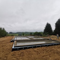 Viflinzider.Bud - Świeżo wylane fundamenty pod budynek, zabezpieczone czarną folią izolacyjną z białymi wypustkami, widoczne wystające pręty zbrojeniowe, w tle krajobraz polny i pochmurne niebo, trzech mężczyzn w tle.