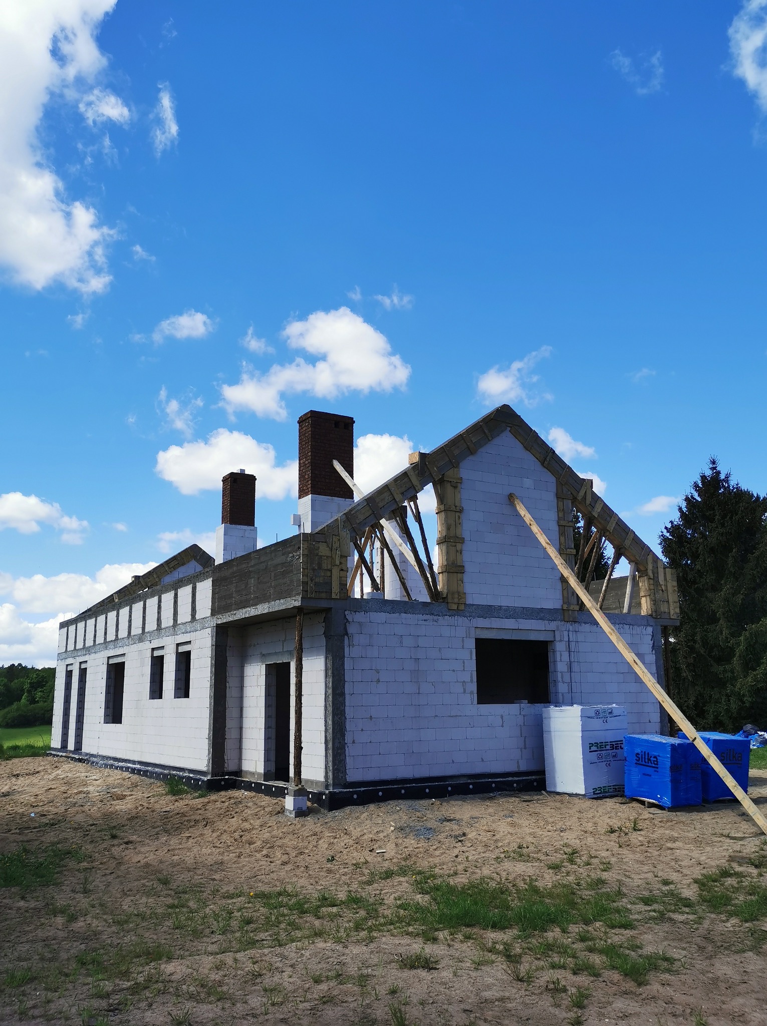 Niedokończony budynek z białych bloczków, widoczna konstrukcja dachu z elementami drewnianymi, dwa kominy z cegły, materiały budowlane Silka na ziemi i błękitne niebo w tle.