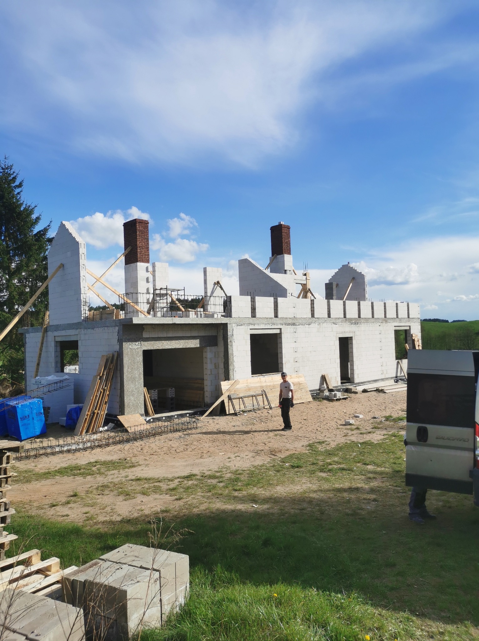 Budowa domu jednorodzinnego z białego bloczka, widoczne kominy z cegły, częściowo zadaszony garaż, teren budowy z materiałami i pracownikiem w oddali, błękitne niebo.