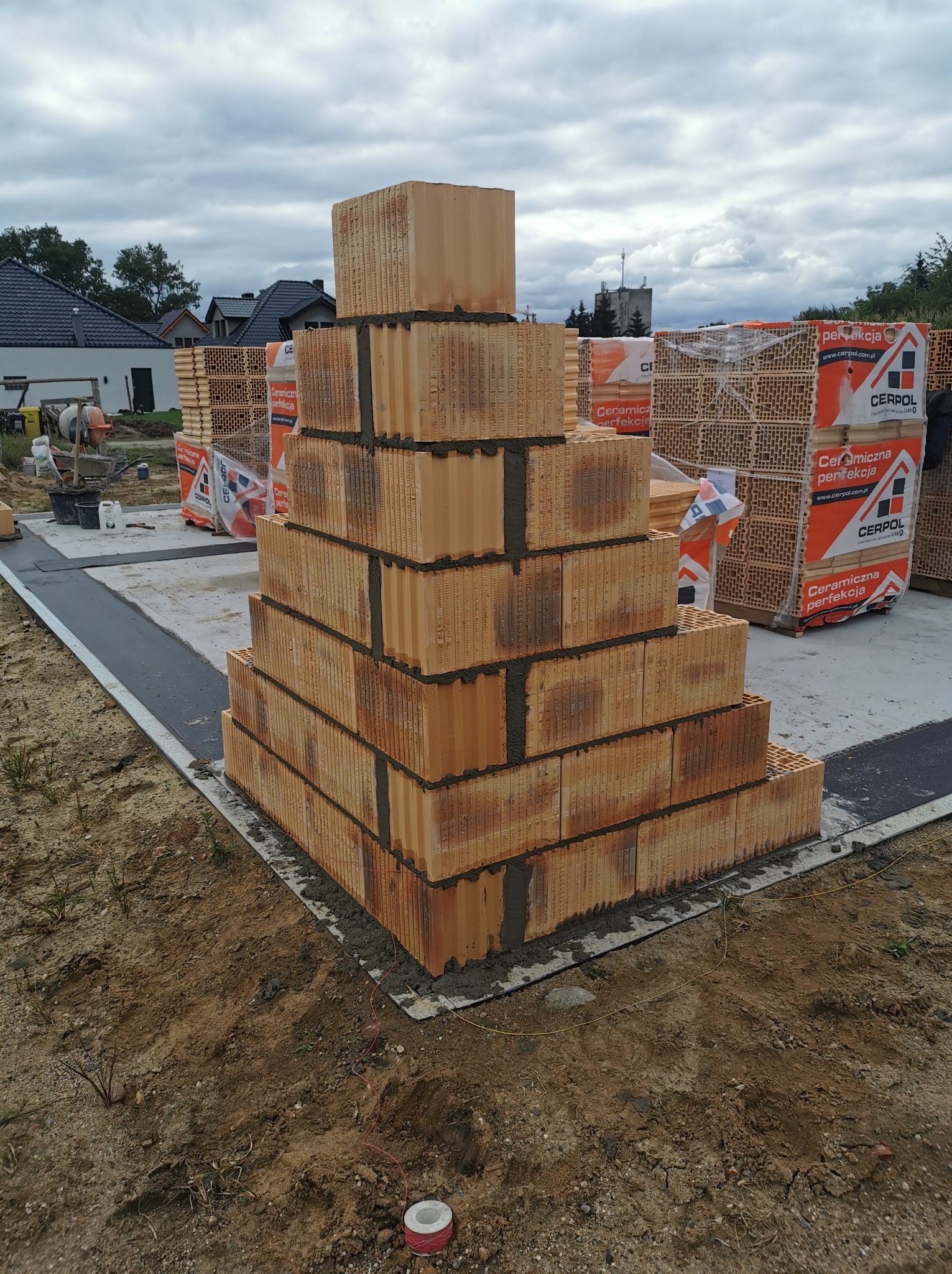 Wznoszenie narożnika domu z bloczków ceramicznych na fundamencie, widoczne palety z bloczkami Cerpol, betonowa podstawa, przygotowany teren budowy, pochmurne niebo.