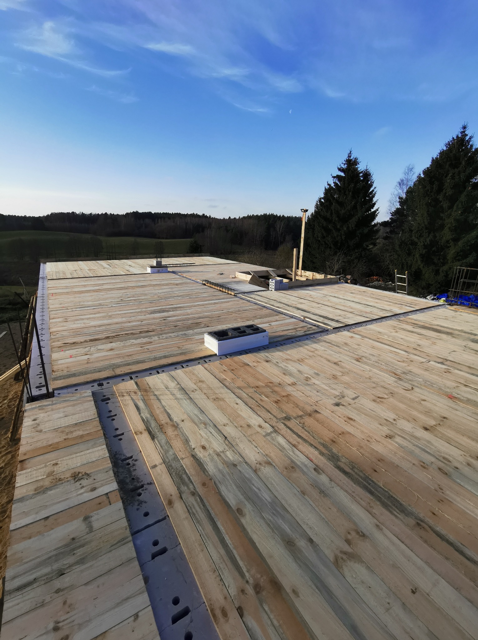 Wczesny etap budowy domu murowanego: widok z góry na drewniany szalunek stropu, z widocznymi elementami konstrukcyjnymi, otworami wentylacyjnymi i zbrojeniami, na tle zielonego krajobrazu...