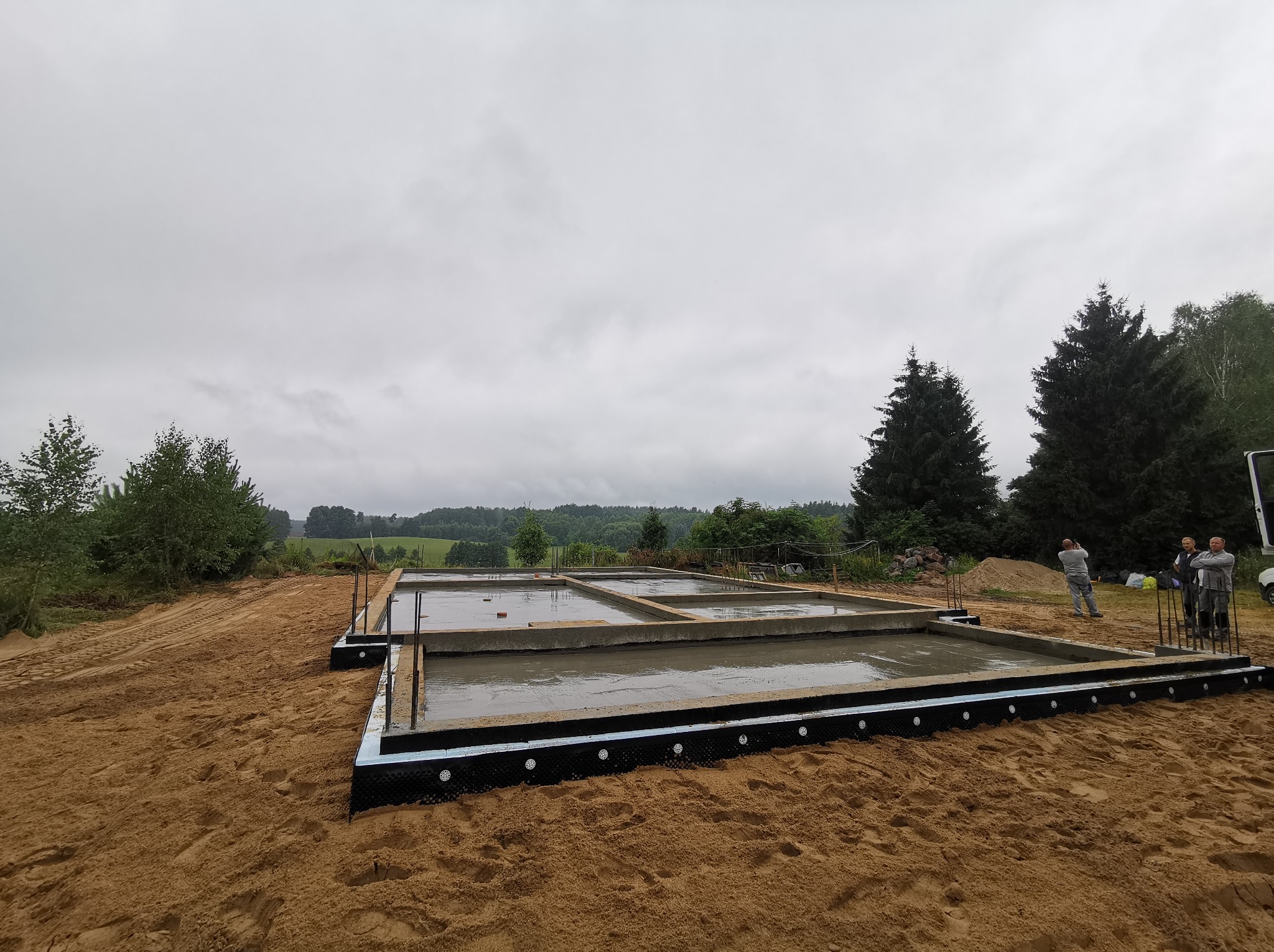 Świeżo wylane fundamenty pod budynek, zabezpieczone czarną folią izolacyjną z białymi wypustkami, widoczne wystające pręty zbrojeniowe, w tle krajobraz polny i pochmurne niebo, trzech mężczyzn w tle.