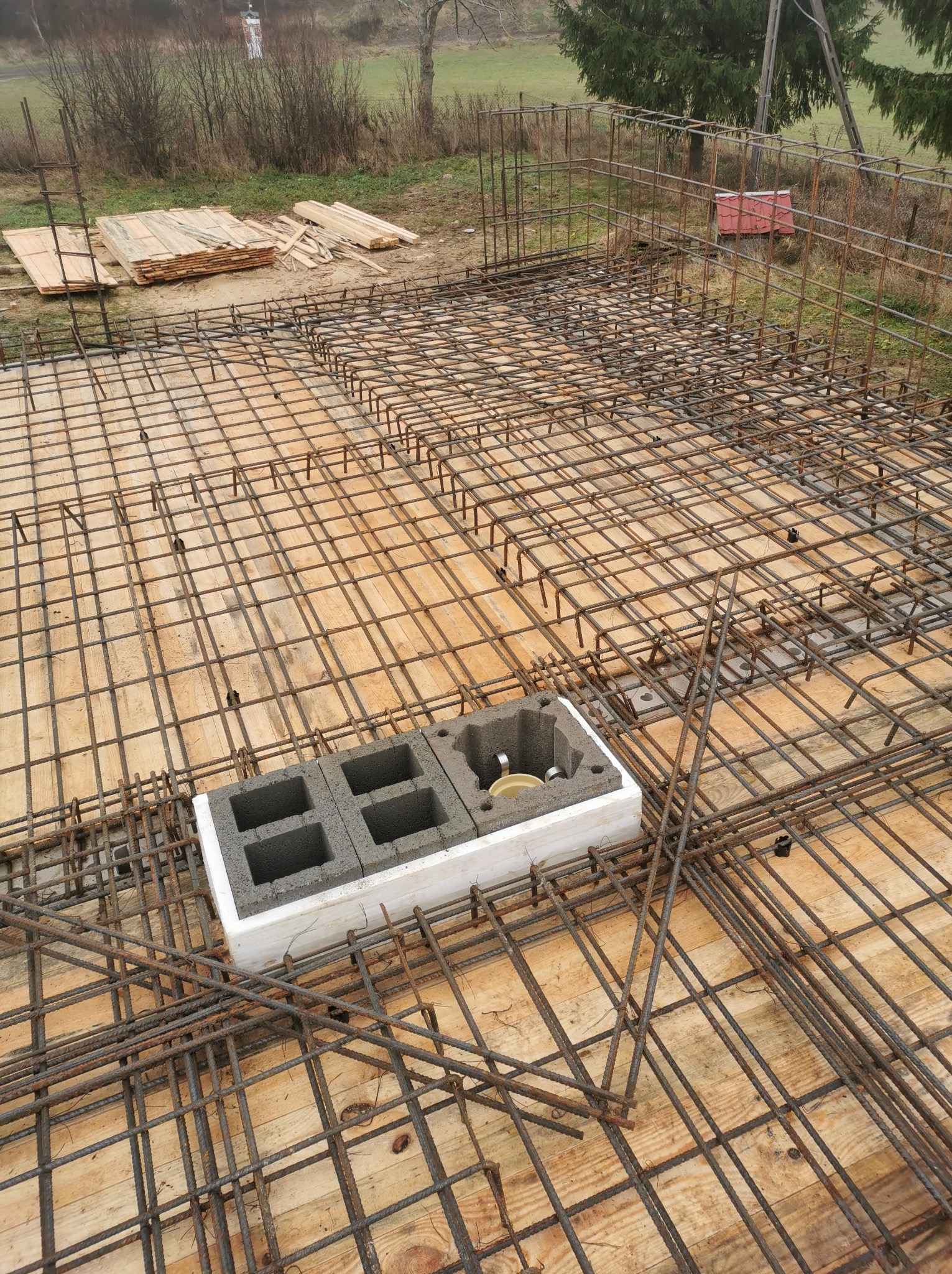 Zbrojenie płyty fundamentowej z widocznymi bloczkami betonowymi i elementami instalacji wewnątrz styropianowej obudowy, przygotowane do zalania betonem.