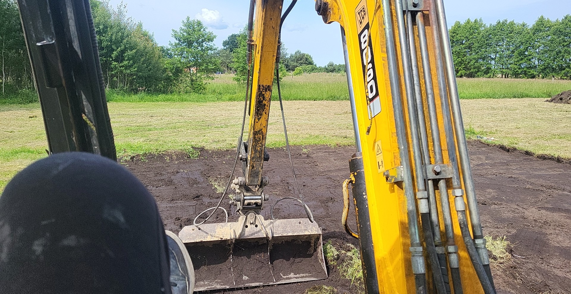 Żółta koparka JCB z łyżką wypełnioną ziemią na tle zielonego trawnika i drzew, widok z kabiny operatora.