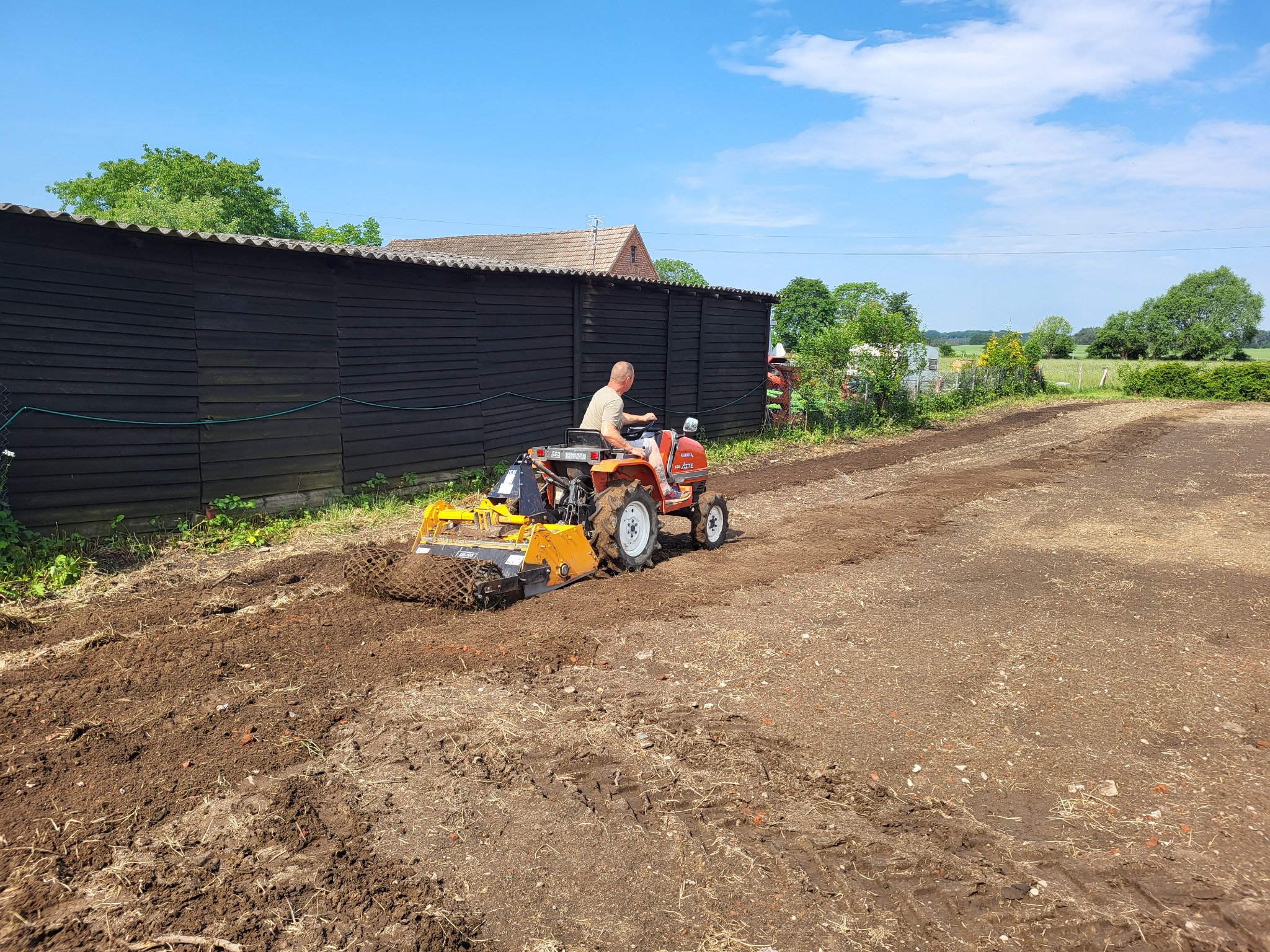 Operator na pomarańczowym traktorze z glebogryzarką wyrównuje ziemię na działce, widoczny czarny drewniany budynek w tle oraz zielone drzewa i błękitne niebo.