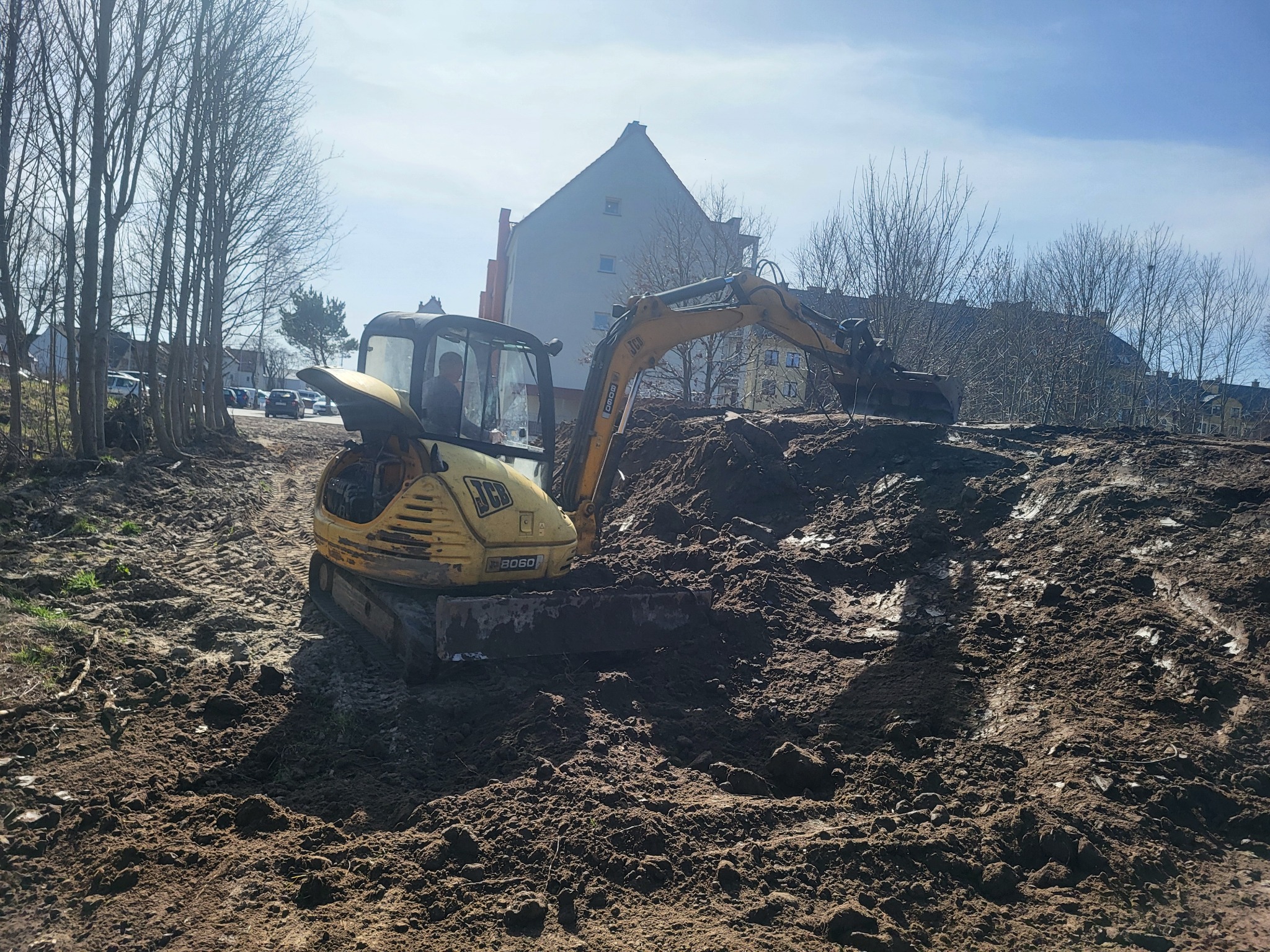 Żółta koparka JCB 8060 na placu budowy, z pryzmą ziemi przed łyżką i budynkami w tle, przy częściowo zachmurzonym niebie.