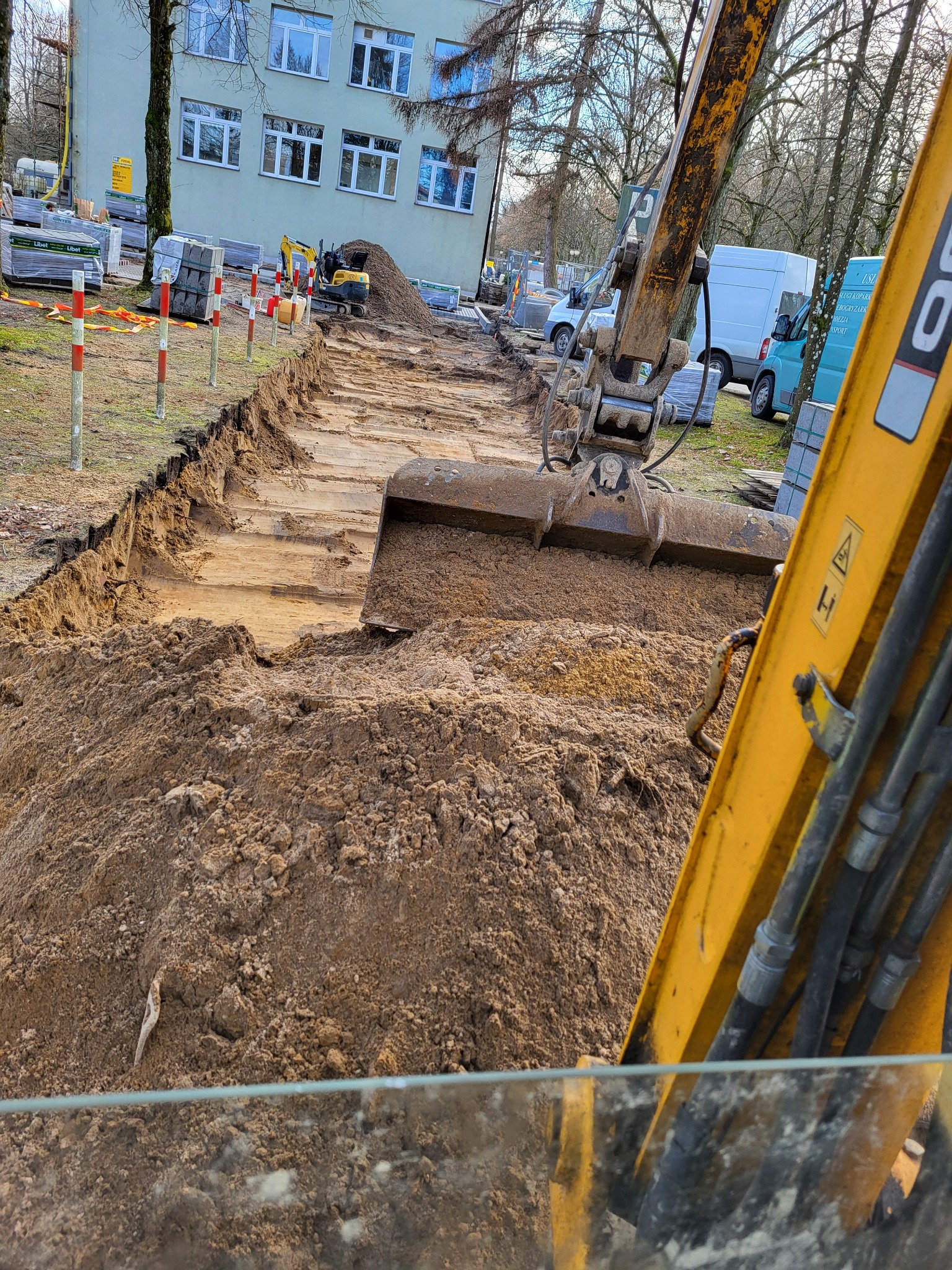 Żółta koparka z łyżką nabiera ziemię, w tle wykopany rów, palety z kostką brukową, budynek i inne pojazdy