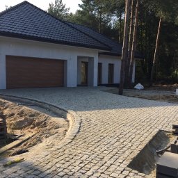 Adaśko firma budowlana - Ułożony z jasnoszarej kostki granitowej podjazd do nowoczesnego domu z brązową bramą garażową, widoczne niezakończone prace przy krawężniku i paleta z kostką.