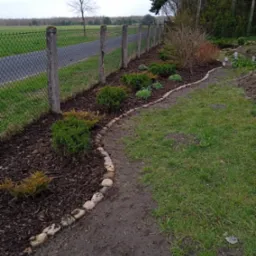 Ułożony brzeg rabaty z kamieni, obsadzona rabata z krzewami iglastymi i kora, w tle ogrodzenie z siatki.
