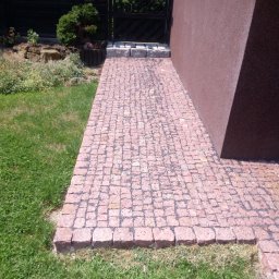 Adaśko firma budowlana - Różowa ścieżka z kostki na trawniku, prowadząca do ciemnej furtki i brązowej ściany budynku.