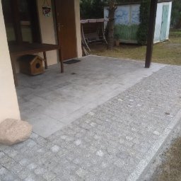 Adaśko firma budowlana - Wyłożony szarym brukiem taras przydomowy z granicą z kostki granitowej, widok na budynek z drewnianym stołem i budką dla ptaków, w tle huśtawka i malowany schowek.