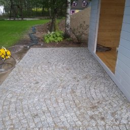 Adaśko firma budowlana - Granitowa nawierzchnia ułożona w łukowaty wzór przed drewnianą altaną w ogrodzie, widoczny fragment trawnika, drzewa i mały domek w tle.