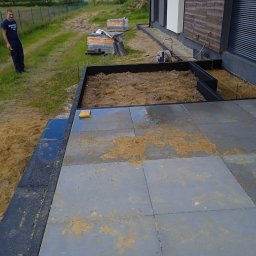 Adaśko firma budowlana - Układanie szarej kostki brukowej na mokro w Łodzi, widoczne przygotowane podłoże, obrzeża i palety z kostką w tle, pracownik nadzoruje postęp prac.