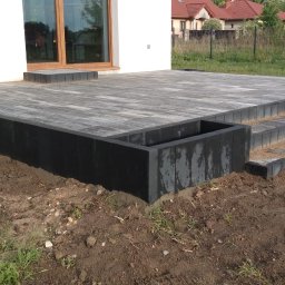Adaśko firma budowlana - Wykończony taras z kostki brukowej w odcieniach szarości, z czarną obudową i schodami, przylegający do białego domu z drewnianą stolarką okienną.