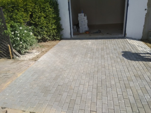 Ułożona szara kostka brukowa prowadząca do garażu, z widoczną roślinnością po bokach i stosem kostki wewnątrz garażu.