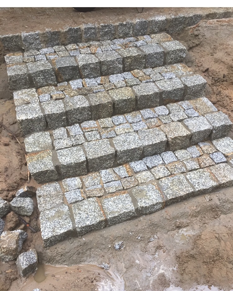 Schody z kostki granitowej w trakcie budowy; widoczne niedociągnięcia w ułożeniu oraz mokre podłoże.