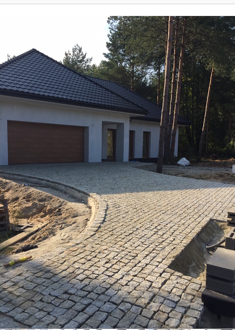 Ułożony z jasnoszarej kostki granitowej podjazd do nowoczesnego domu z brązową bramą garażową, widoczne niezakończone prace przy krawężniku i paleta z kostką.