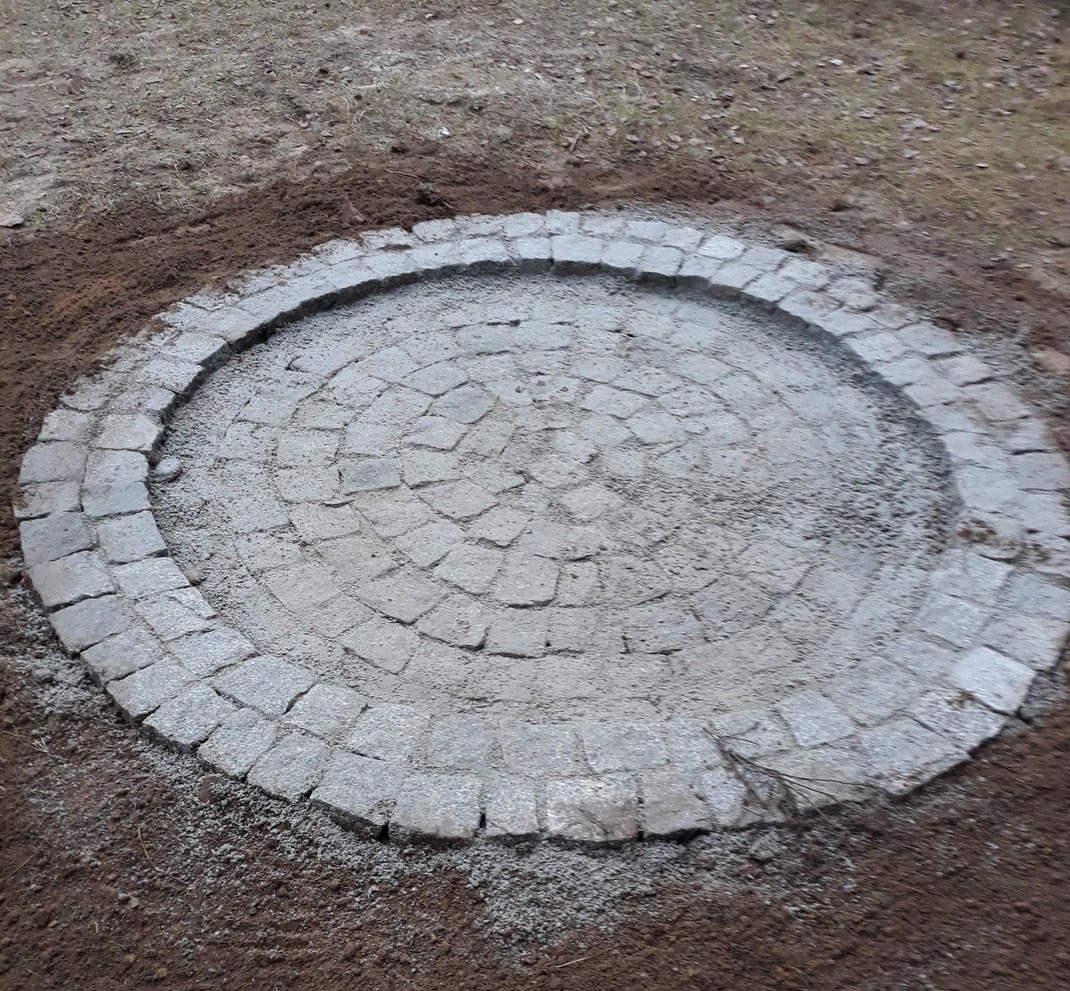 Okrągła powierzchnia wyłożona szarą kostką brukową w trakcie prac, otoczona ziemią, widoczne niedociągnięcia w ułożeniu i drobne zanieczyszczenia.