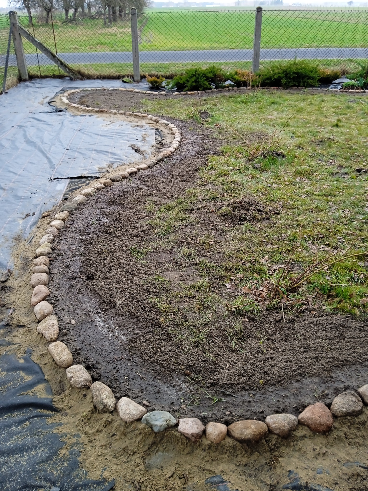 Kręta ścieżka w ogrodzie w trakcie tworzenia, wyznaczona kamieniami, obok czarna agrowłóknina i trawnik z kępami trawy, w tle siatka ogrodzeniowa.