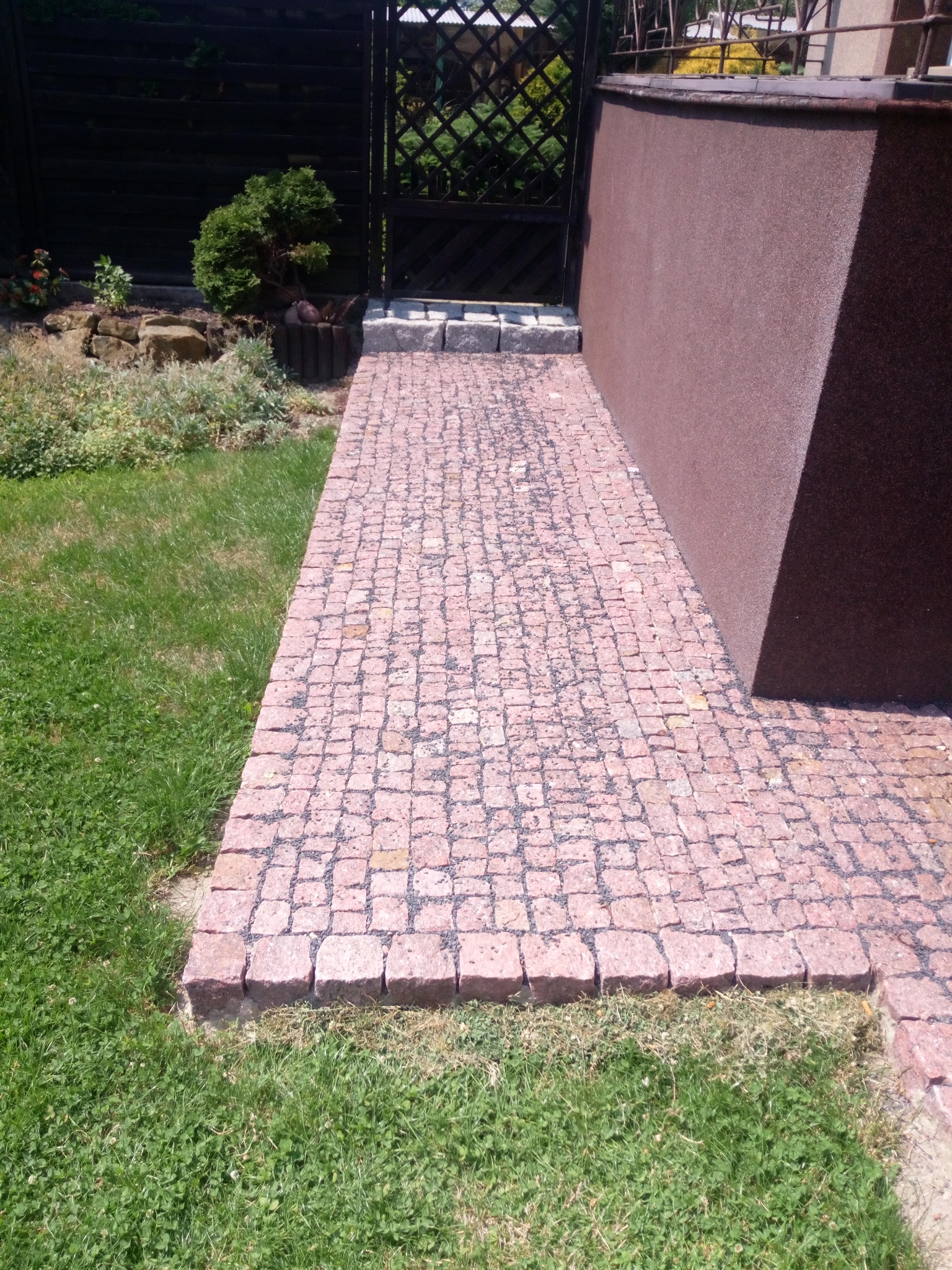Różowa ścieżka z kostki na trawniku, prowadząca do ciemnej furtki i brązowej ściany budynku.