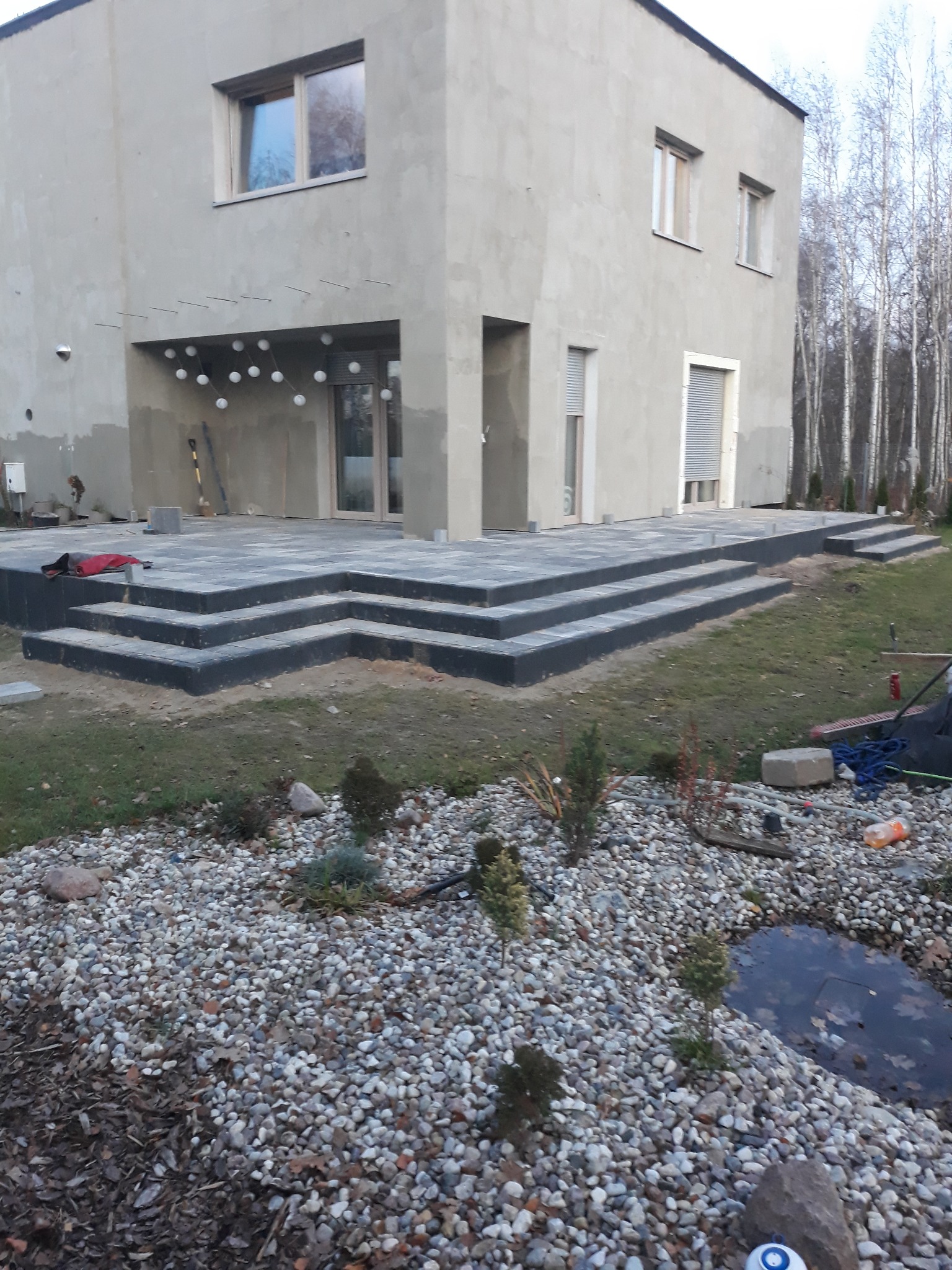 Nowoczesny dom z szarego tynku z tarasem i schodami z ciemnej kostki brukowej, otoczony ogrodem z kamieniami i małym oczkiem wodnym.