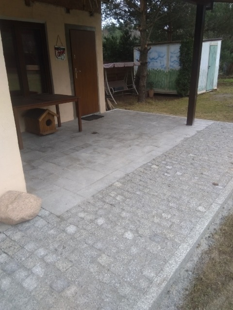 Wyłożony szarym brukiem taras przydomowy z granicą z kostki granitowej, widok na budynek z drewnianym stołem i budką dla ptaków, w tle huśtawka i malowany schowek.