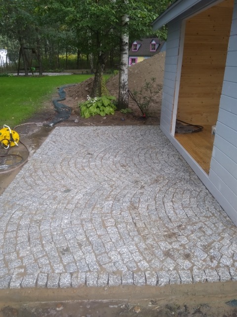 Granitowa nawierzchnia ułożona w łukowaty wzór przed drewnianą altaną w ogrodzie, widoczny fragment trawnika, drzewa i mały domek w tle.