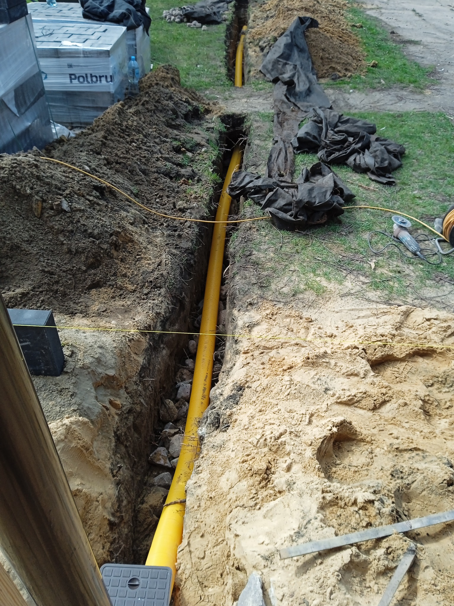 Wąski wykop w ziemi z żółtą rurą karbowaną na dnie, widoczne kamienie, obok hałda ziemi i materiały budowlane, w tle trawa i narzędzia.
