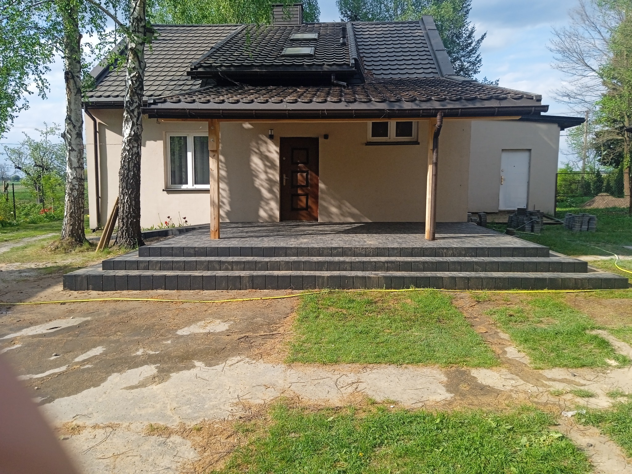 Schody wejściowe do domu wyłożone szarą kostką brukową, widok na elewację, brązowe drzwi wejściowe i fragment ogrodu z żółtym wężem ogrodowym.