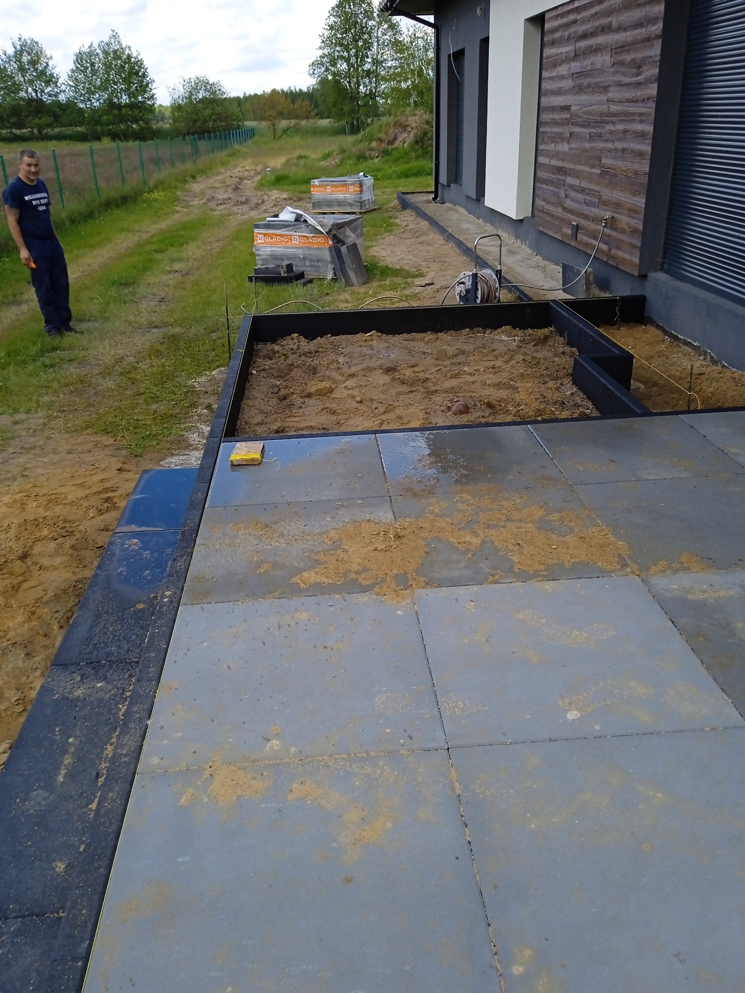 Układanie szarej kostki brukowej na mokro w Łodzi, widoczne przygotowane podłoże, obrzeża i palety z kostką w tle, pracownik nadzoruje postęp prac.