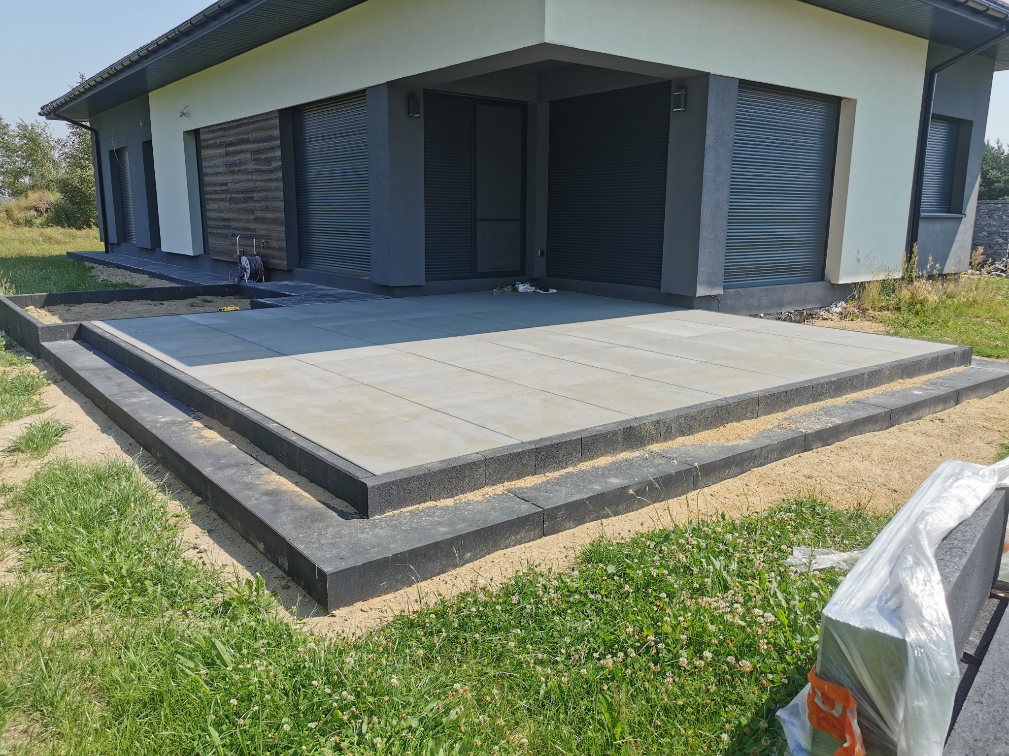 Nowoczesny taras z jasnych płyt betonowych z czarną obwódką, prowadzący do domu z szarymi roletami zewnętrznymi, widoczne ślady piasku na trawie.