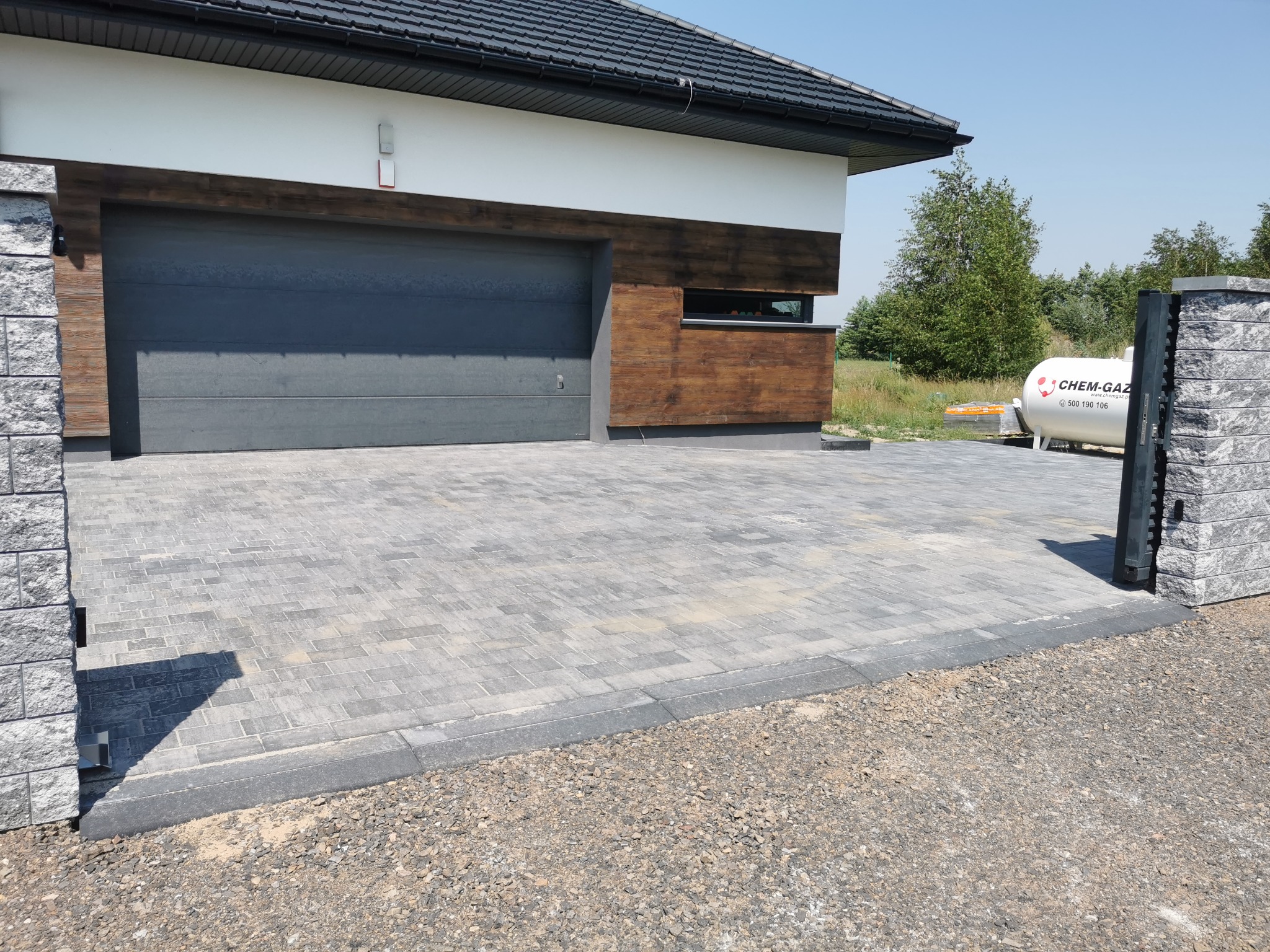 Ułożona szara kostka brukowa przed nowoczesnym domem z garażem i bramą wjazdową, widoczny zbiornik na gaz.