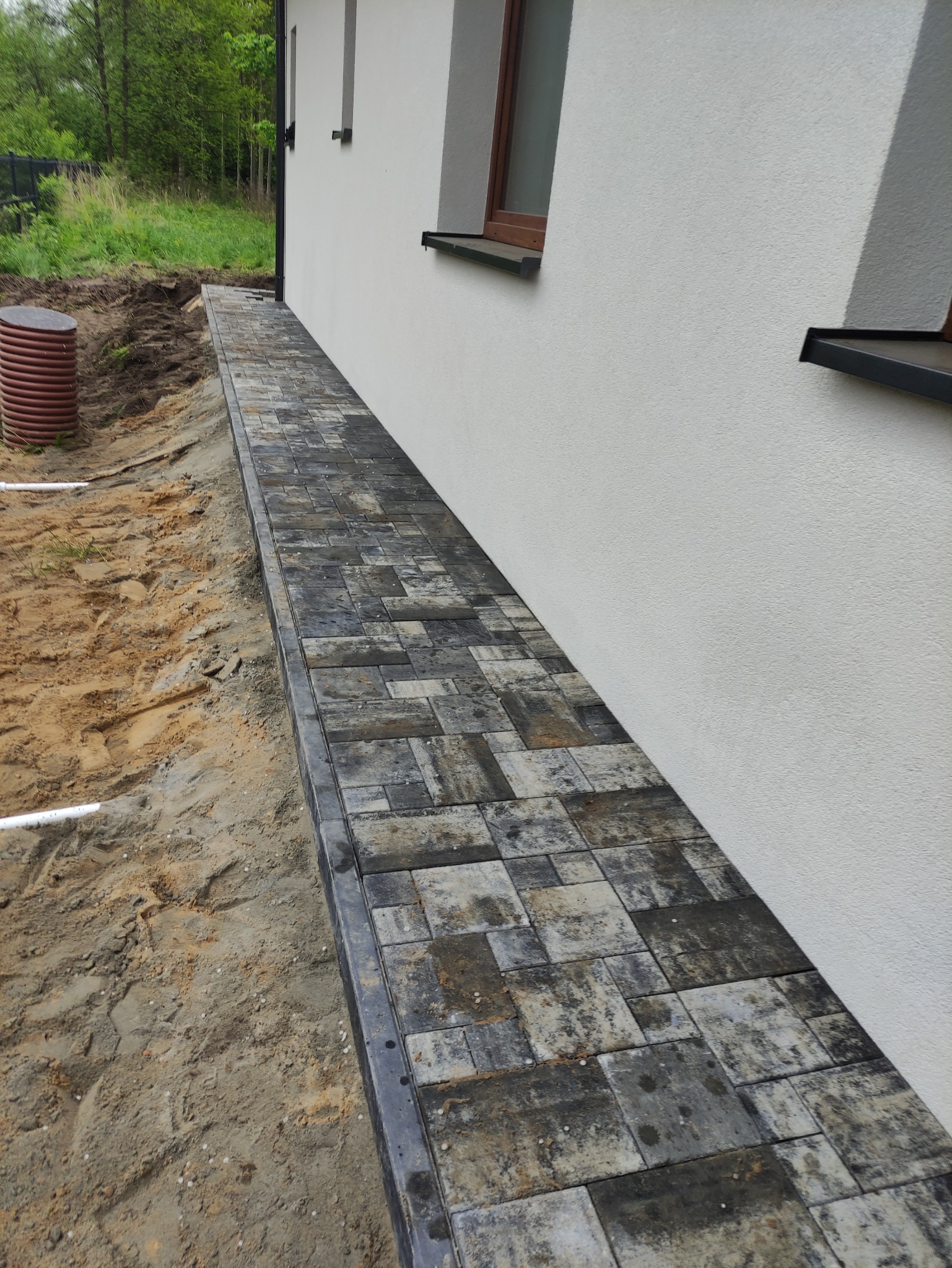 Ścieżka z kostki brukowej w odcieniach szarości i beżu, ułożona wzdłuż białej ściany budynku, widoczny brązowy zbiornik na wodę i ziemia przygotowana pod ogród.