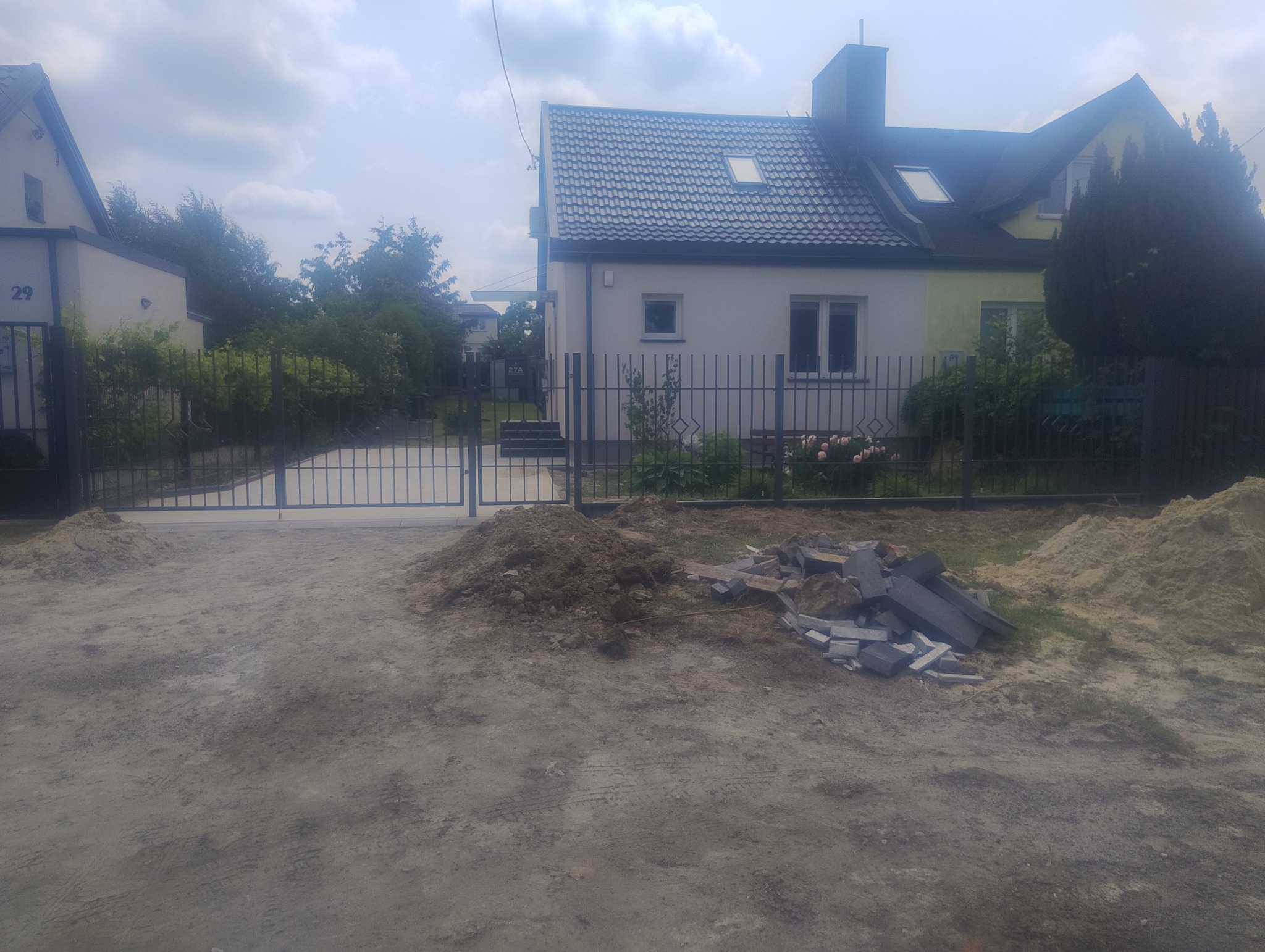 Nowoczesna, grafitowa brama wjazdowa i ogrodzenie panelowe przed domem z szarą dachówką. Na pierwszym planie piasek i kostka brukowa.