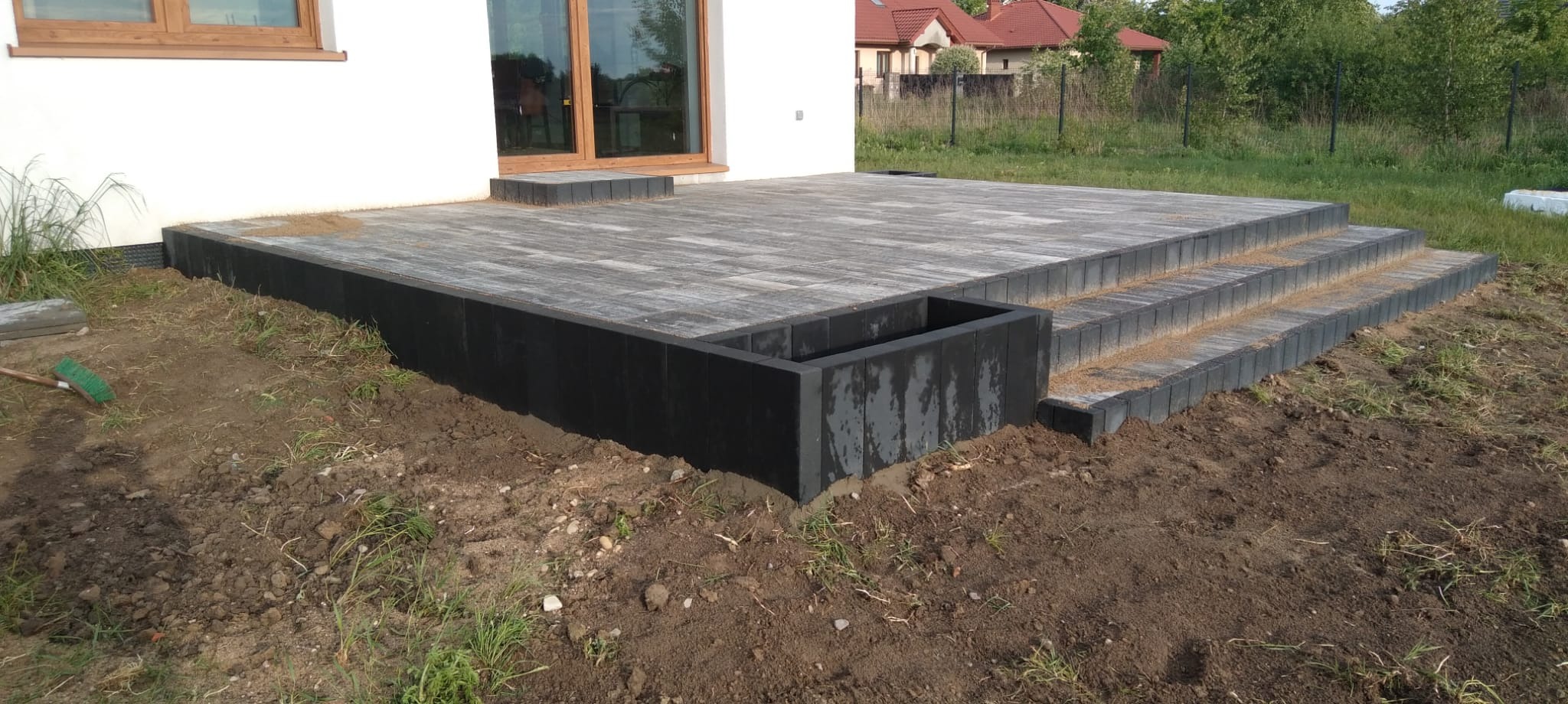 Wykończony taras z kostki brukowej w odcieniach szarości, z czarną obudową i schodami, przylegający do białego domu z drewnianą stolarką okienną.