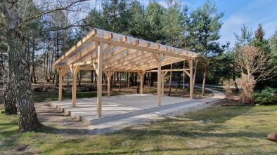 Drewniana wiata z przezroczystym zadaszeniem, osadzona na betonowej platformie z niskimi schodami, otoczona drzewami w słoneczny dzień. Widok na ścieżkę w tle.