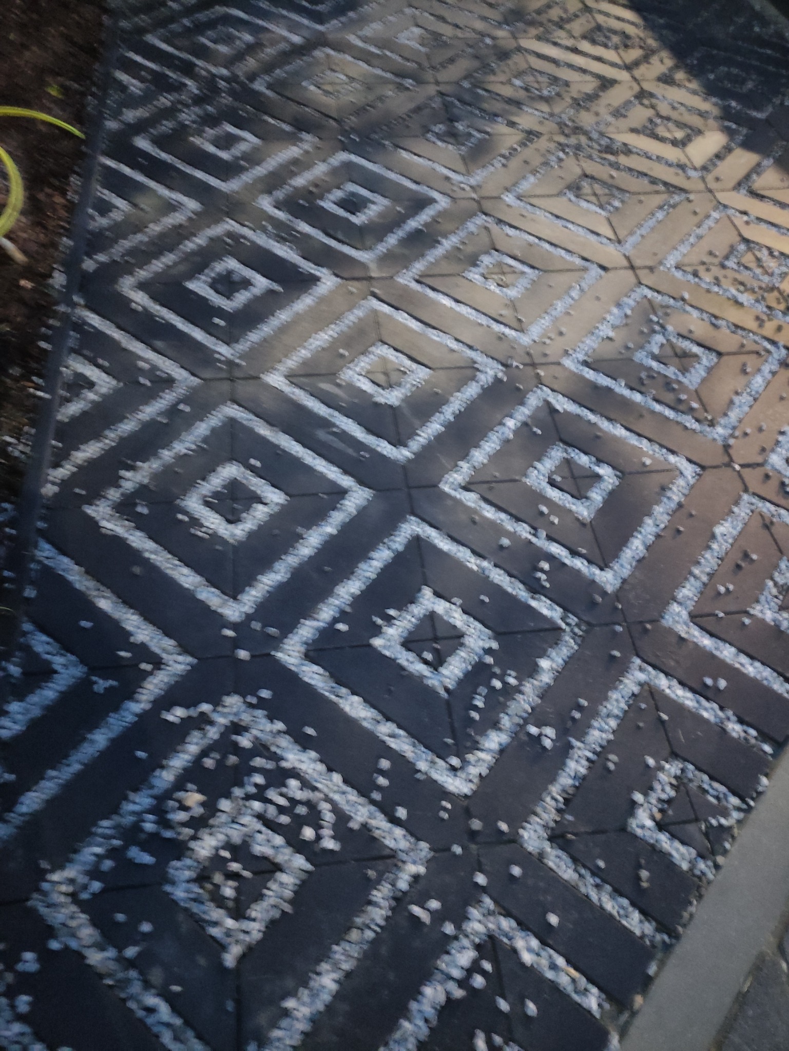 Ciemna kostka brukowa ułożona we wzór rombów, wypełniona drobnymi kamyczkami, tworząc unikalną i dekoracyjną nawierzchnię.