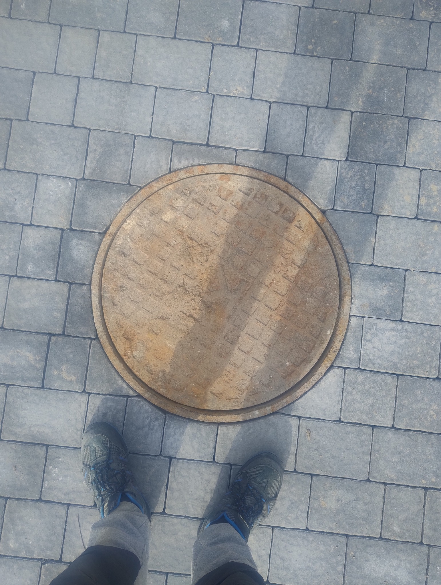 Szara kostka brukowa ułożona wokół okrągłej, żeliwnej studzienki kanalizacyjnej. Widoczne stopy osoby stojącej na kostce.