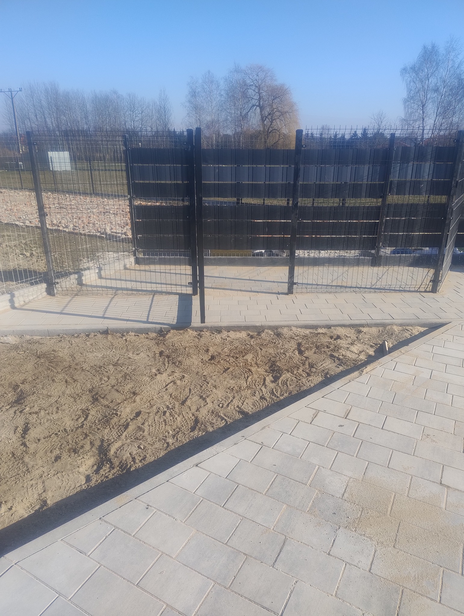 Nowoczesne ogrodzenie panelowe w kolorze grafitowym, z widoczną kostką brukową i piaskiem, sugerujące prace wykończeniowe na posesji.