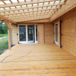 Drewniany taras z zadaszeniem i oknami, wykończony w jasnym odcieniu drewna, z widocznymi detalami zdobniczymi na krawędzi dachu i czarnymi okuciami okiennymi.