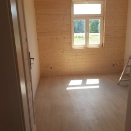 Fazbud - Wnętrze pokoju z drewnianą boazerią ścienną, jasną podłogą, oknem z widokiem na zieleń i fragmentem drabiny w rogu, widoczne przewody elektryczne przy podłodze.