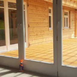 Widok przez białe, przesuwne drzwi balkonowe na drewniany taras i elewację domu z drewnianych bali; na podłodze wewnątrz stoi butelka środka czyszczącego z pomarańczowym rozpylaczem.