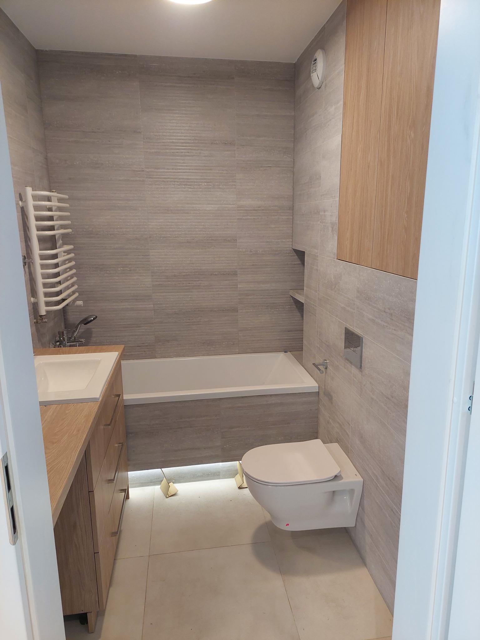 Nowoczesna łazienka z szarymi płytkami imitującymi drewno, podwieszana toaleta, szafka pod umywalką z drewnianym frontem i grzejnik drabinkowy.