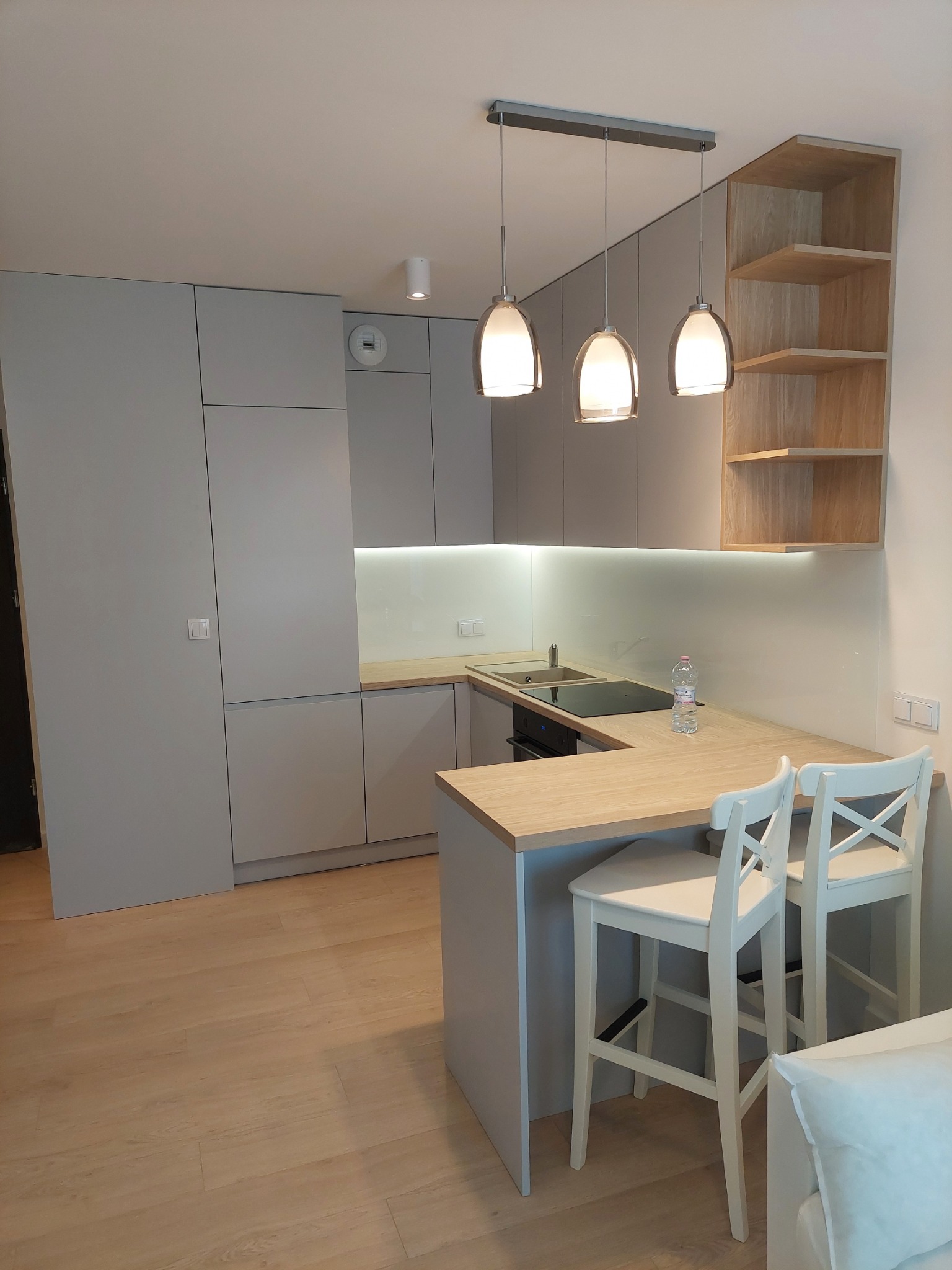 Nowoczesna kuchnia w szarościach z drewnianymi akcentami, oświetleniem podszafkowym LED i trzema wiszącymi lampami nad blatem z dwoma białymi krzesłami barowymi.
